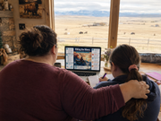 Two people sitting together in a room with a large window view of a field and mountains.